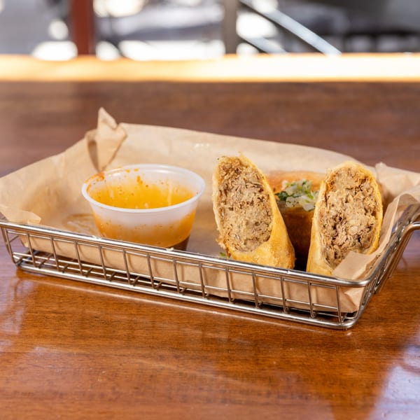 Crispy egg rolls filled with meat, served in a metal basket with a side of dipping sauce.
