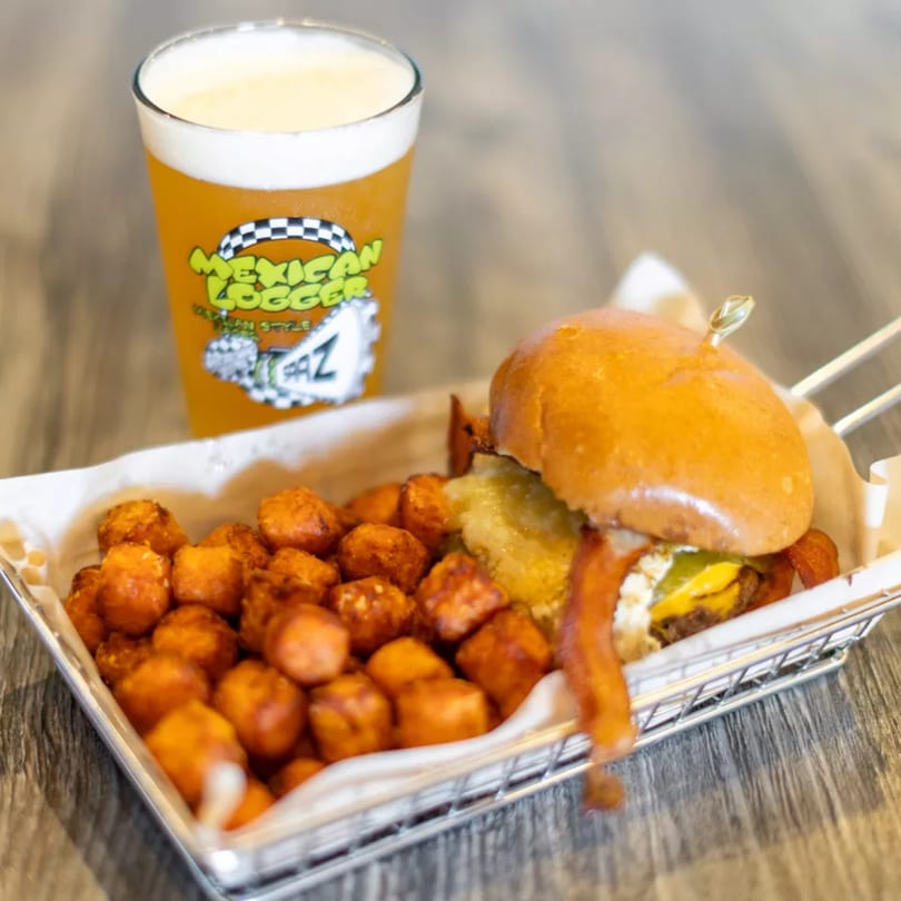 A burger with bacon and cheese, a side of tater tots, and a Mexican Logger beer.