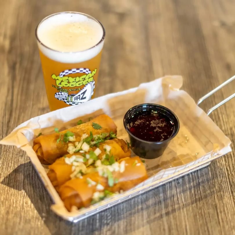 Fried rolls topped with green onions and dipping sauce, next to a glass of Mexican Lager beer.