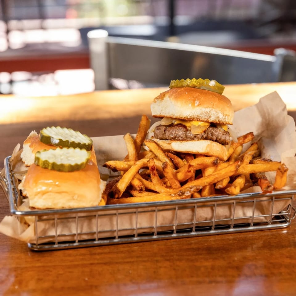 Two Brews sliders with pickles and a side of golden fries in a metal basket on a wooden table.