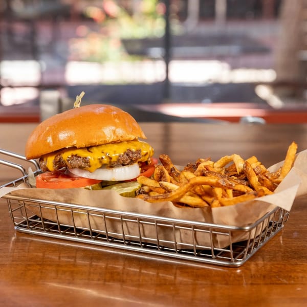 A Brews gourmet burger with melted cheese, tomato, onion, and pickles, served with fries in a metal basket.
