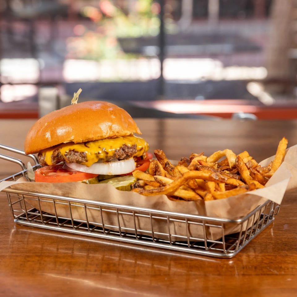 A Brews gourmet burger with melted cheese, tomato, onion, and pickles, served with fries in a metal basket.
