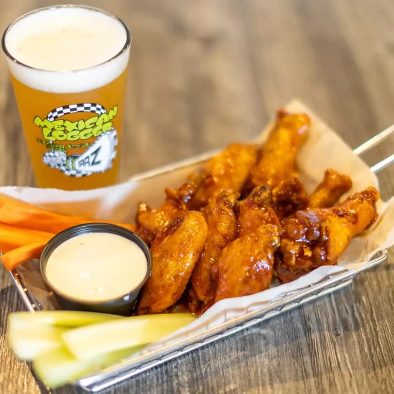 Glazed chicken wings in a basket with dip, carrots, celery, and a glass of Mexican Logger beer.